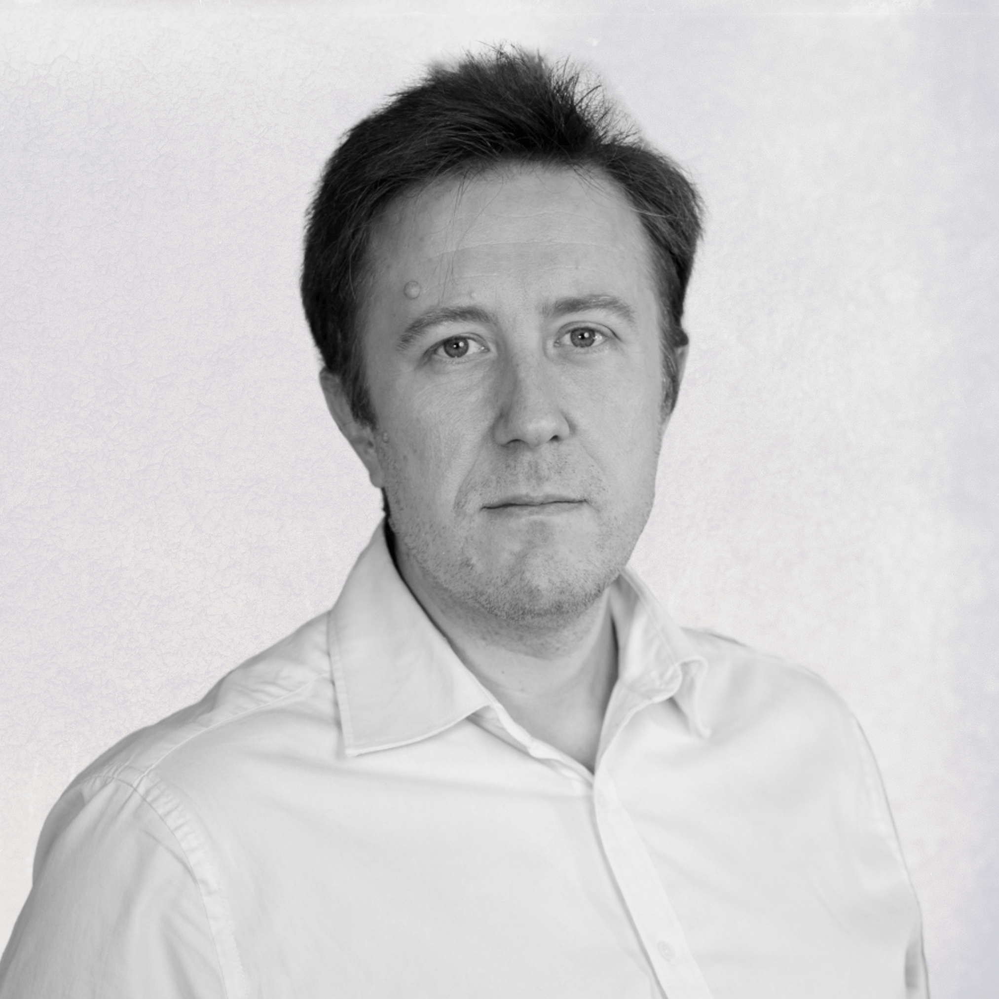 Black and white portrait of a middle-aged man with short dark hair wearing a white collared shirt, looking directly at the camera against a plain light background.