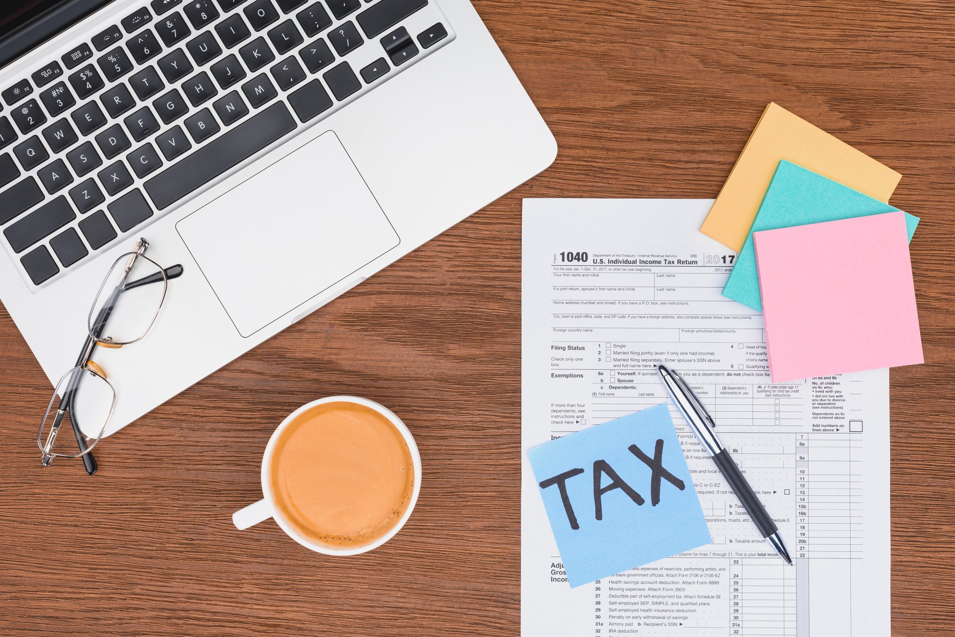 Top view of a laptop keyboard, eyeglasses, a cup of coffee, and a 1040 U.S. tax form with colorful sticky notes and a pen on a wooden desk.
