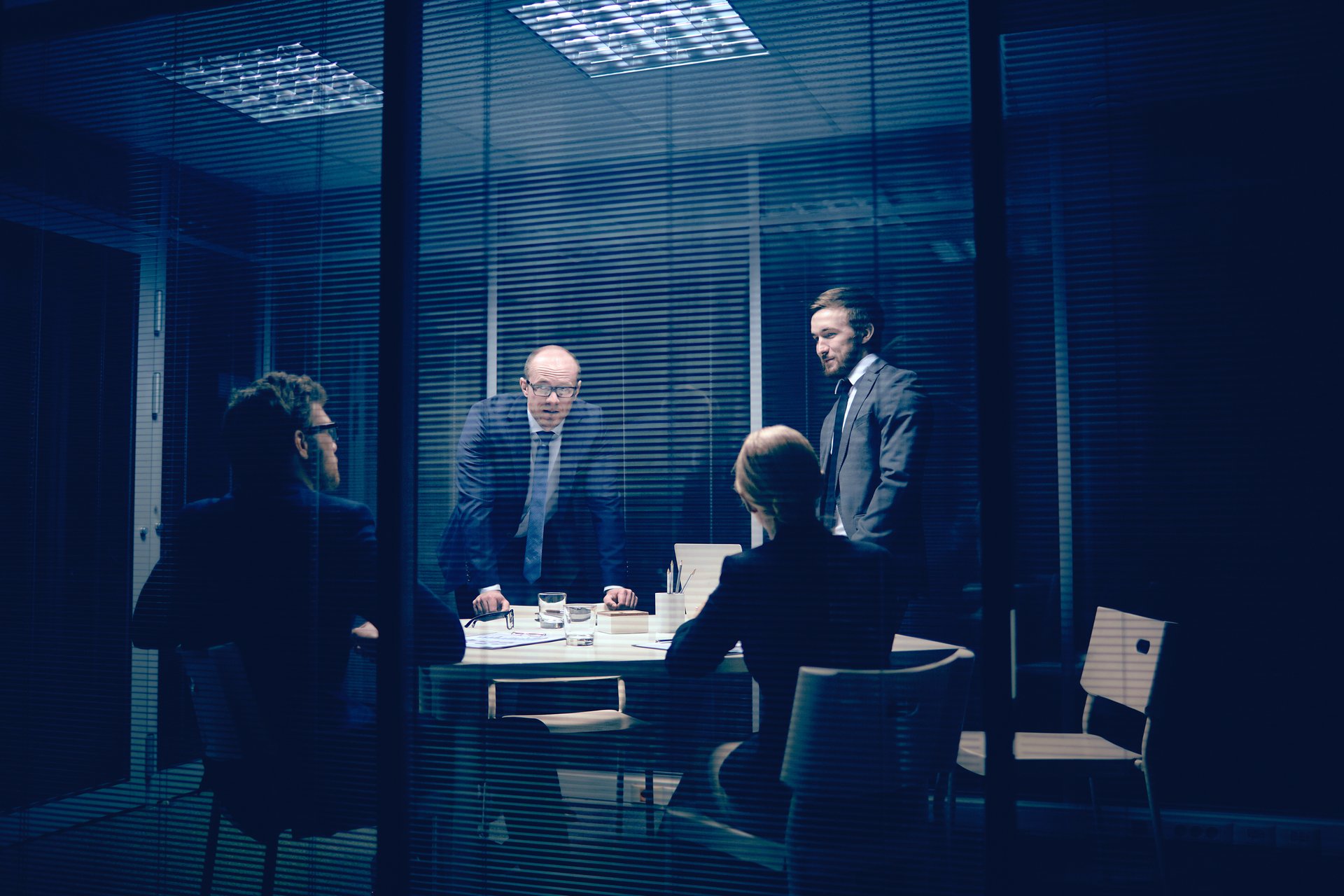 Four business professionals in suits having a meeting around a conference table in a modern office at night.
