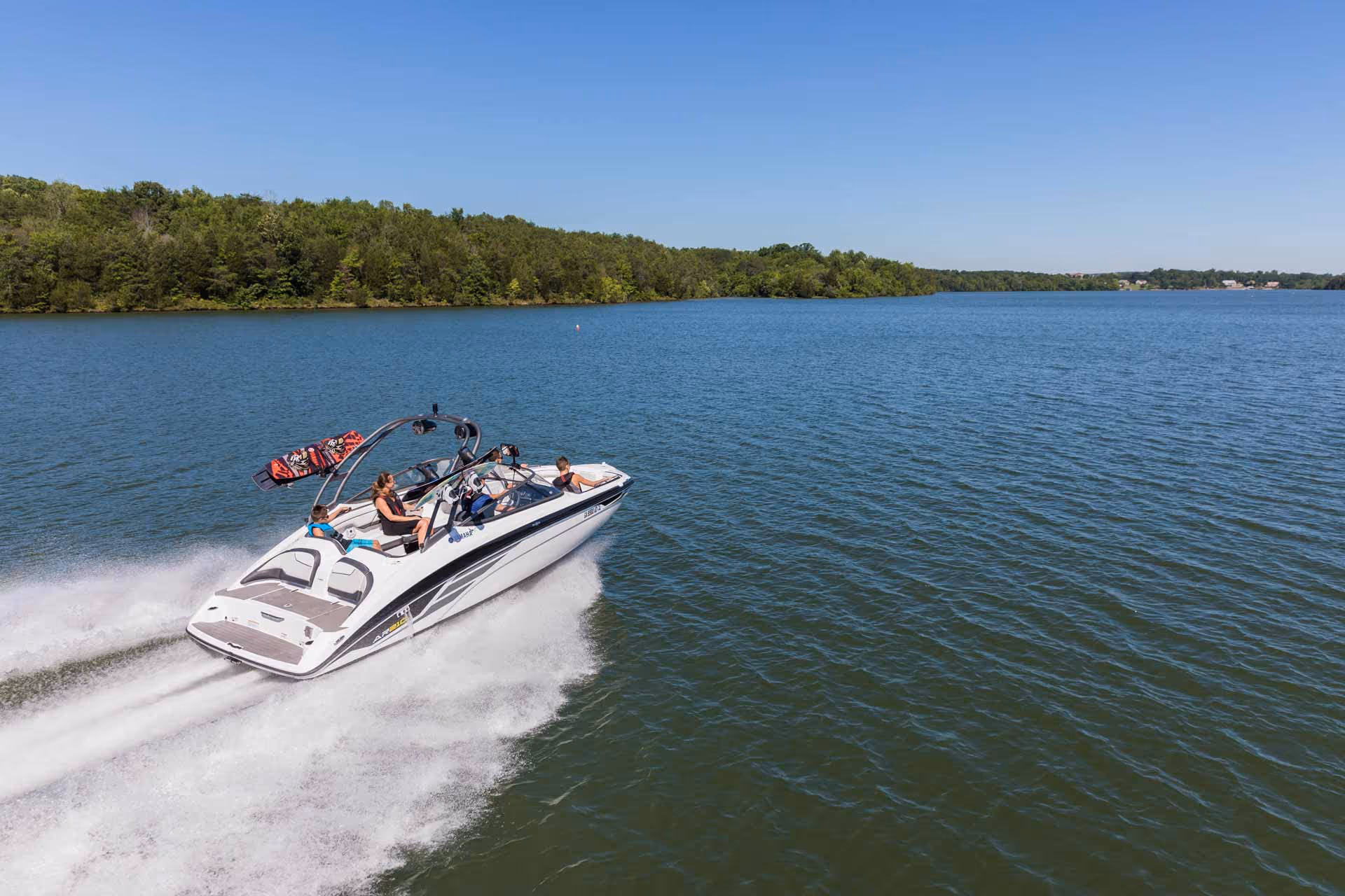 Speedboat with four people cruising on a lake under a clear blue sky near a green forested shore.