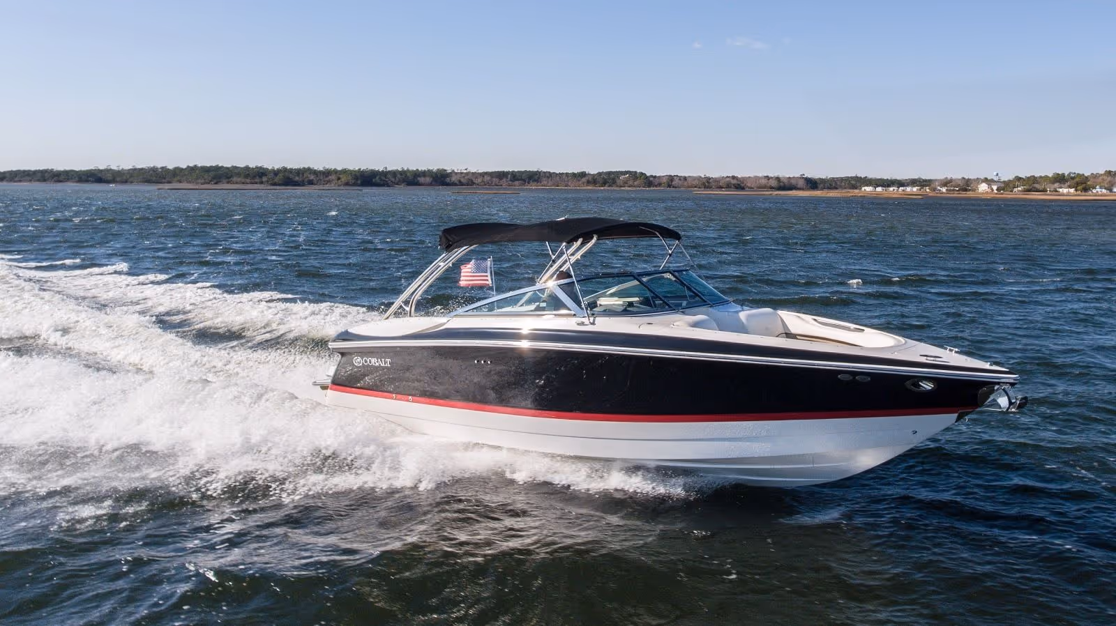Black and white Cobalt speedboat with red stripe cruising on a lake under clear blue sky.