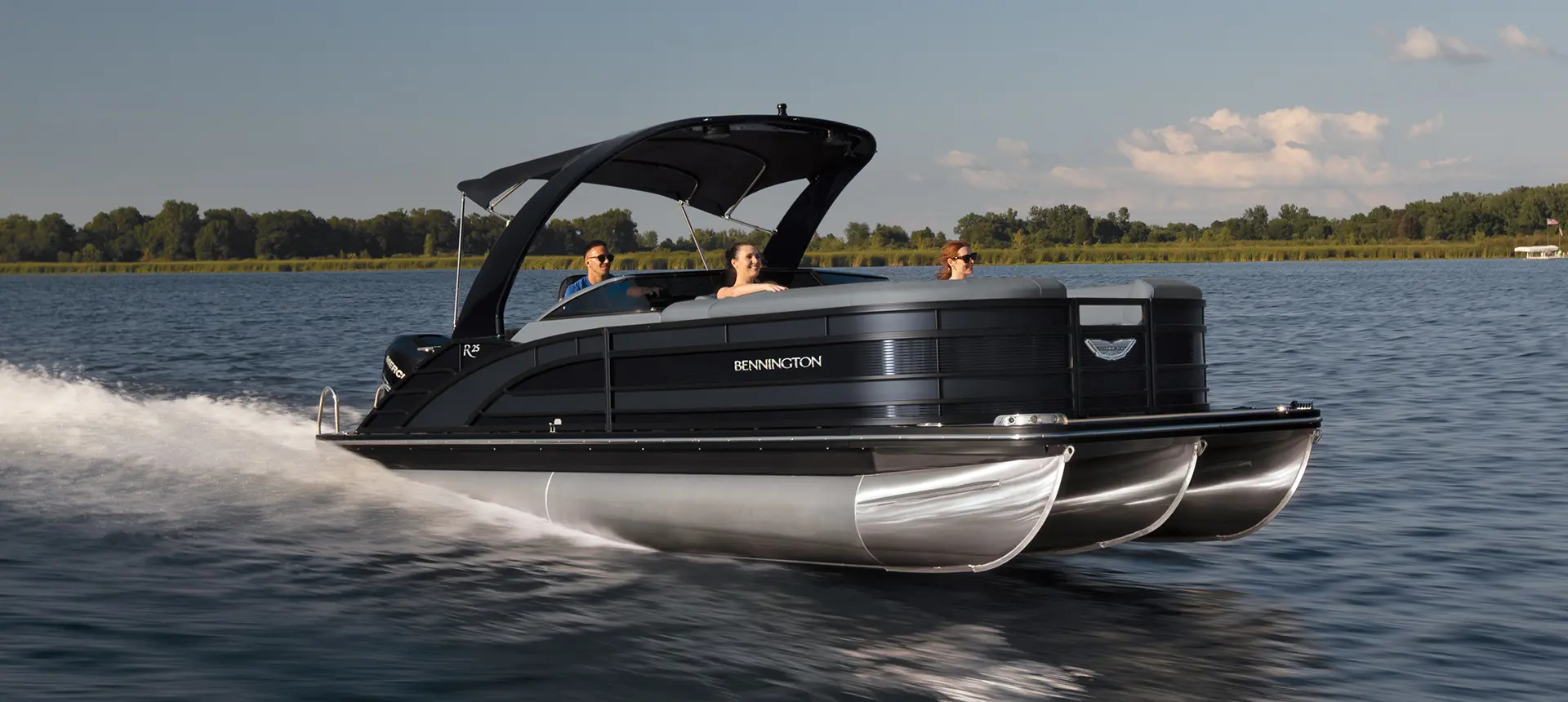 Three people riding a black Bennington pontoon boat on a calm lake with trees in the background.