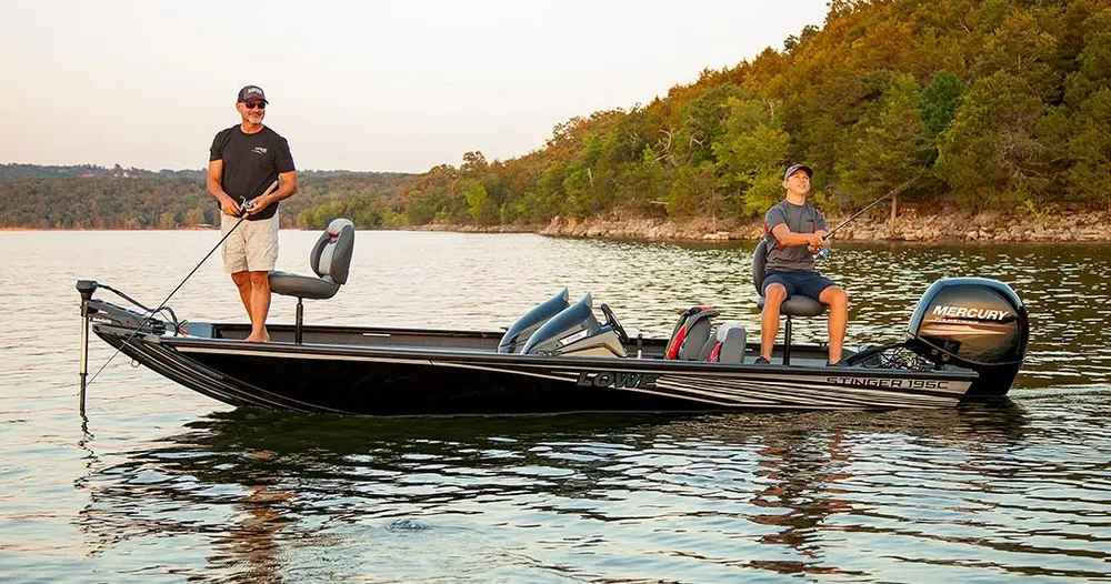 Two men fishing on a black Lowe fishing boat on a calm lake surrounded by forested hills.