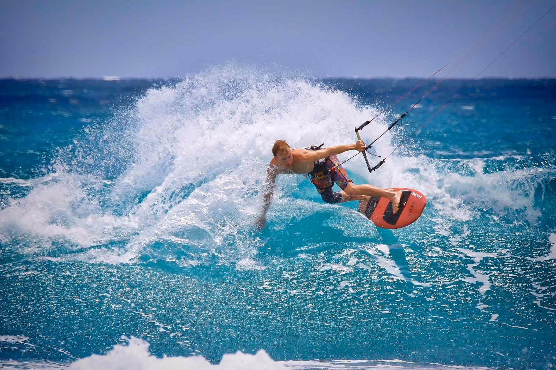 Kite surfer in colorful shorts riding a wave on a bright red board in blue ocean water.