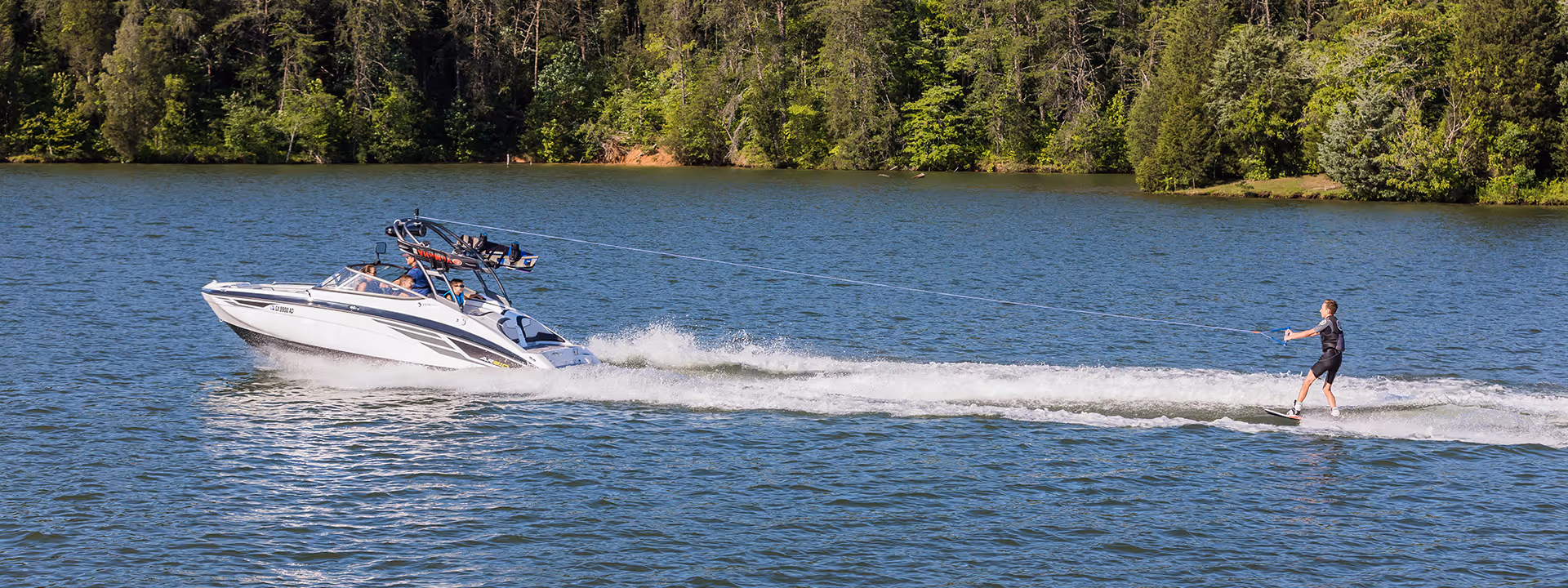 Person waterskiing on a lake being pulled by a white motorboat with passengers and trees in the background.