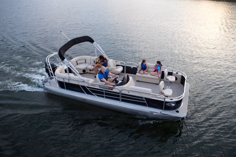 Four people wearing life jackets riding on a black and beige pontoon boat on calm water.