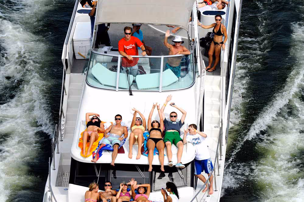 Group of people sunbathing and relaxing on a moving yacht over water with wake trails.