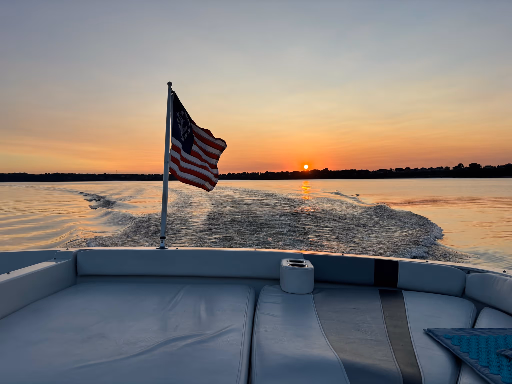 American flag on boat with sun setting over calm lake and shoreline in the distance.