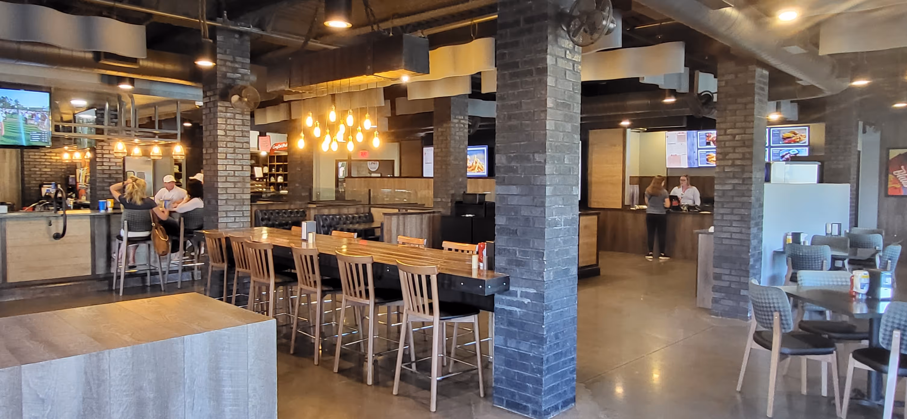 Modern bar and grill interior with wood tables, high chairs, patrons, and hanging lights.