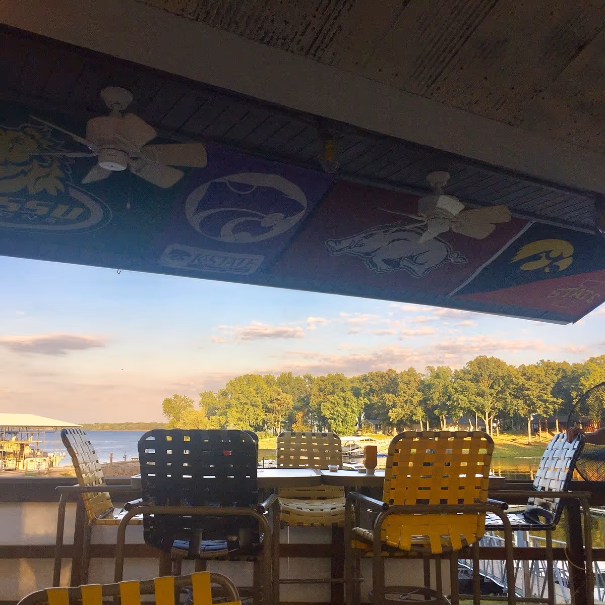Outdoor seating area with colorful chairs overlooking a lake and trees under a ceiling with sports team banners.