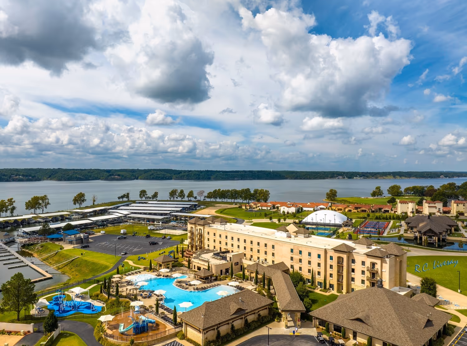 Aerial view of lakeside resort with pools, tennis courts, marina, and buildings under a cloudy blue sky.