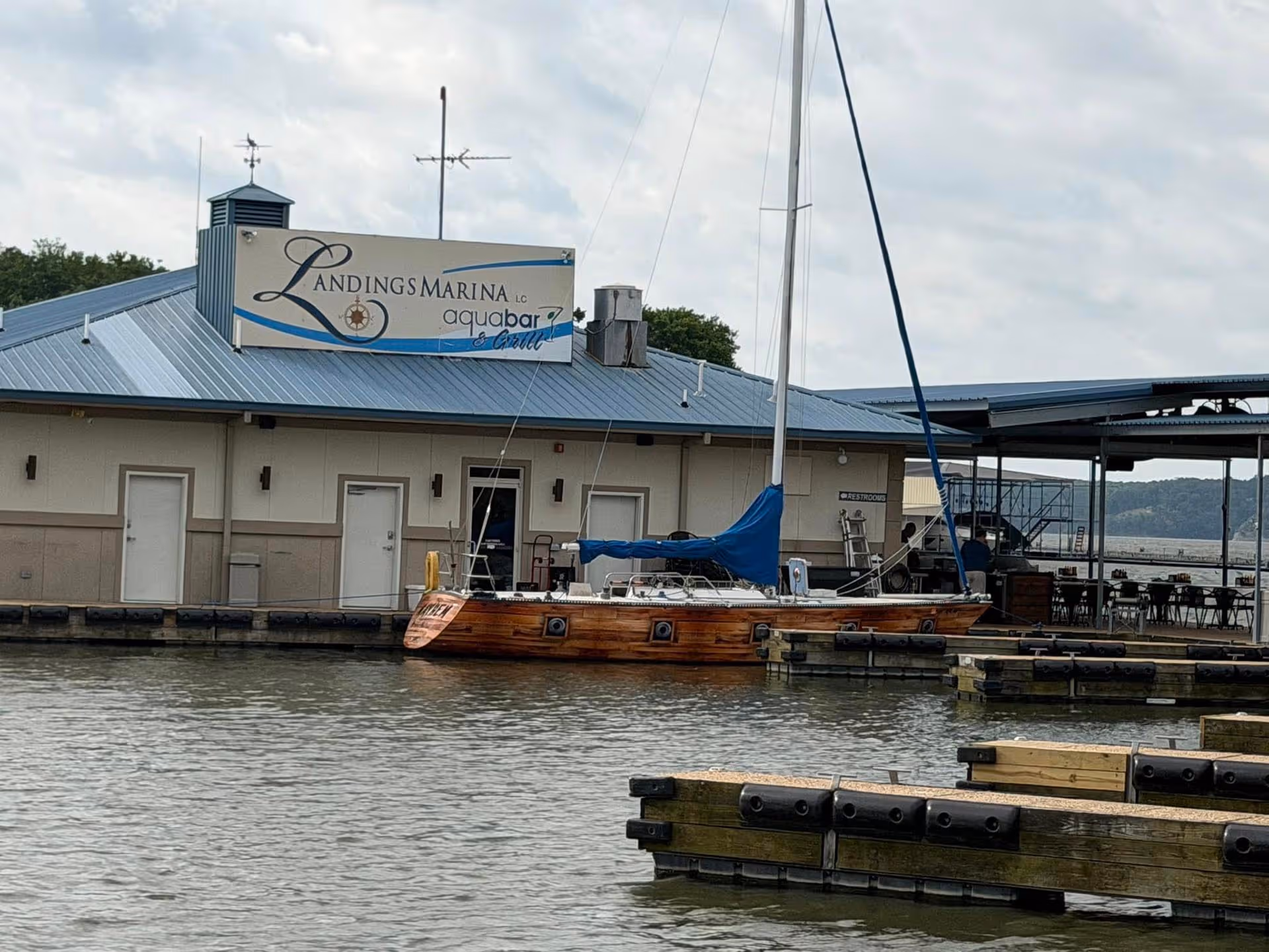 Wooden sailboat docked at Landings Marina aquabar and grill building under cloudy sky.