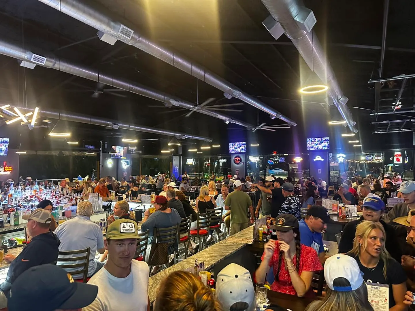 Crowded bar and restaurant with people sitting at tables and the bar, illuminated with ceiling lights.
