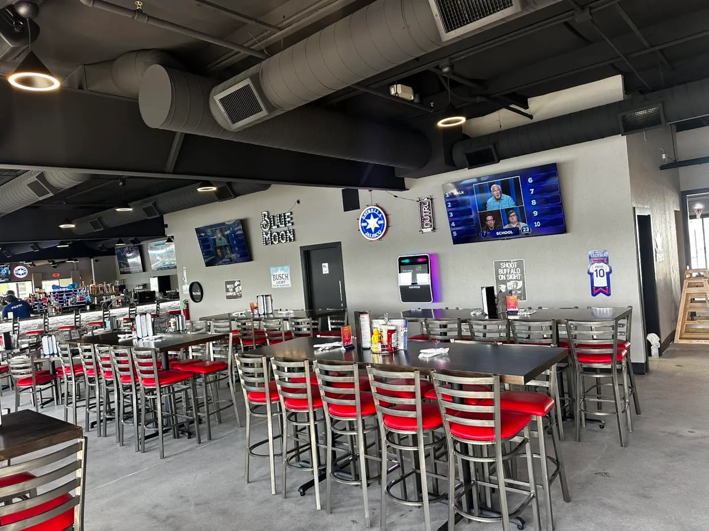 Interior of a modern bar with high tables and red cushioned chairs, TVs on walls, and visible ductwork.