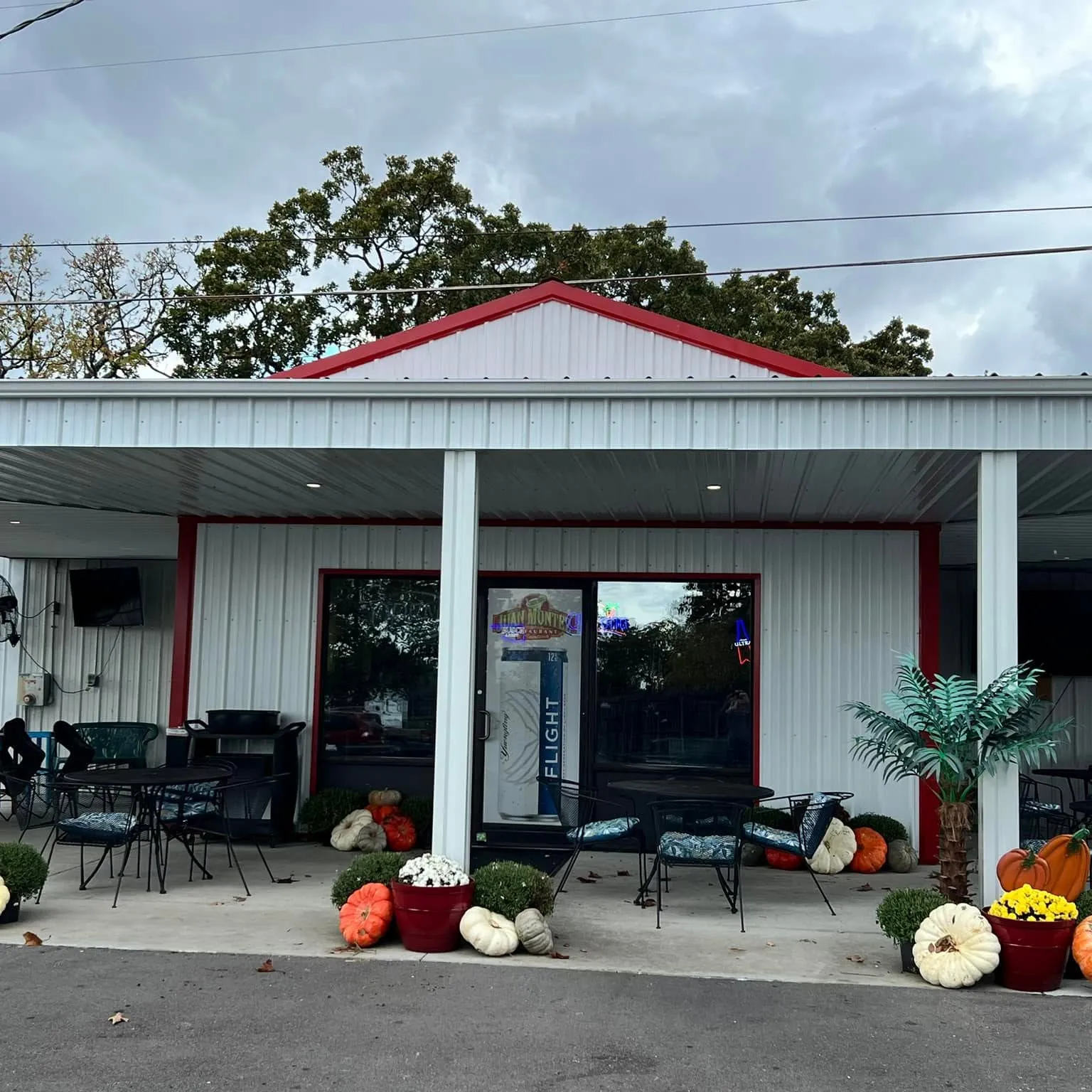 Outdoor seating area with black tables, chairs, pumpkins, and potted plants outside a white and red building.