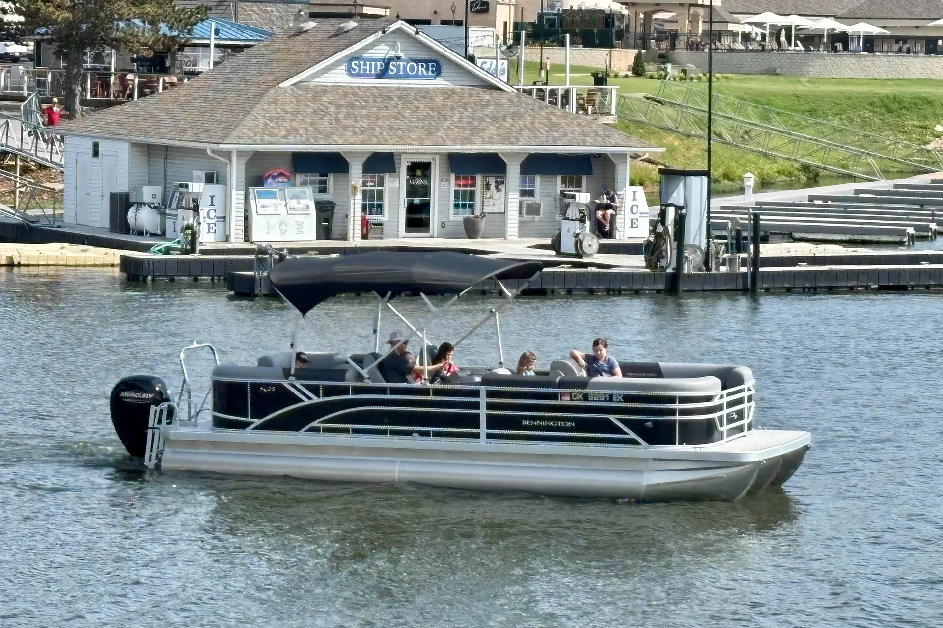 Five people riding on a black Bennington pontoon boat near a dock with a Ship Store building.