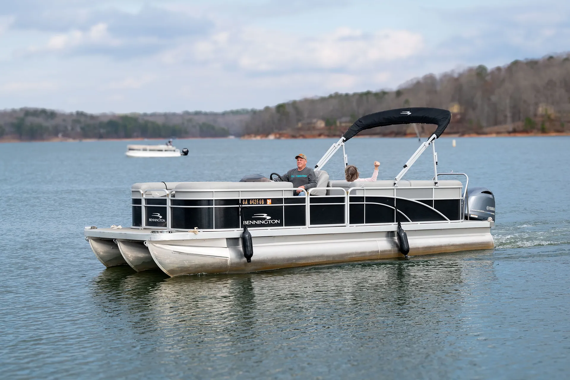 Two people on a black Bennington pontoon boat cruising on a lake with trees in the background.