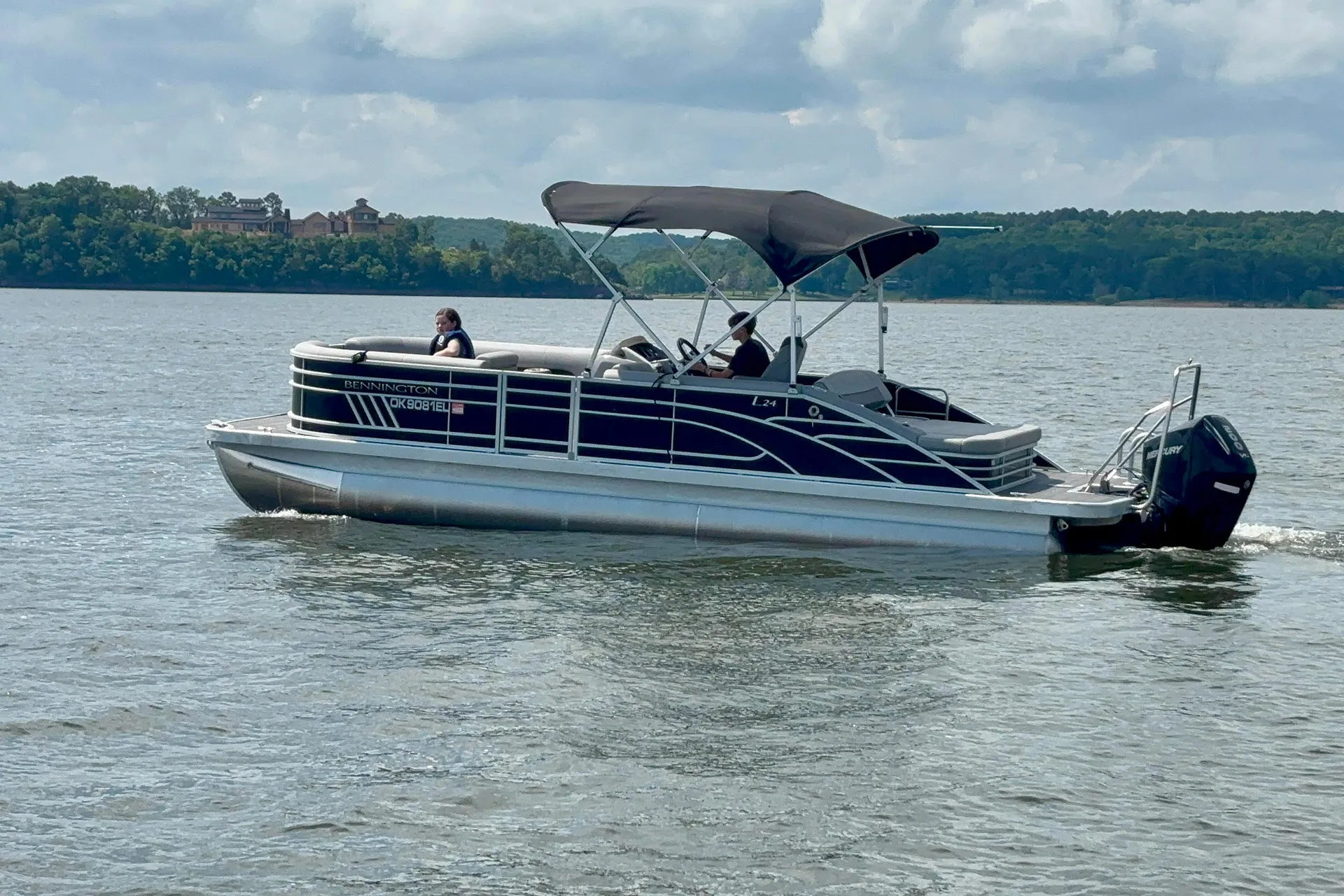 Pontoon boat with two passengers cruising on a calm lake under a cloudy sky.