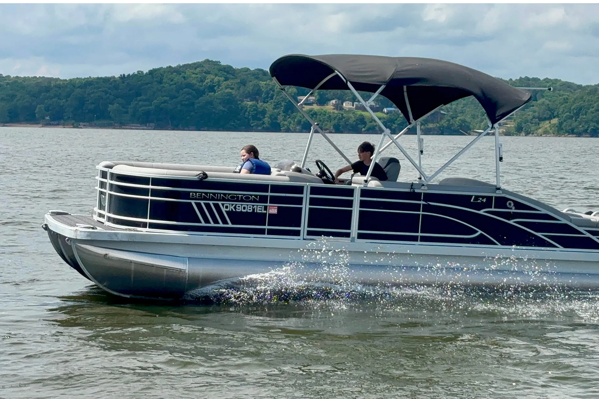 Two people riding a black Bennington pontoon boat on a lake with tree-covered hills in the background.