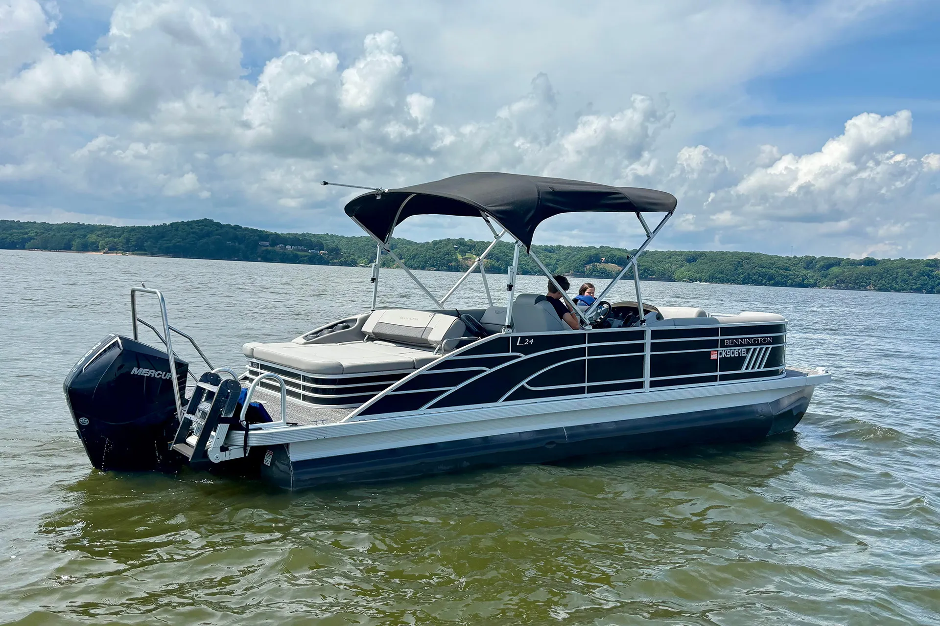 Black and gray Bennington pontoon boat with two people on a lake under a partly cloudy sky.