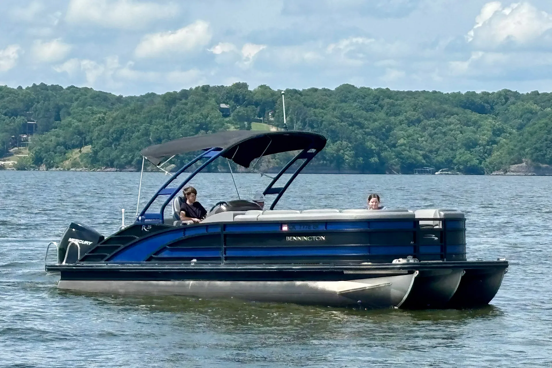 Blue Bennington pontoon boat with two people cruising on a lake surrounded by forested hills.