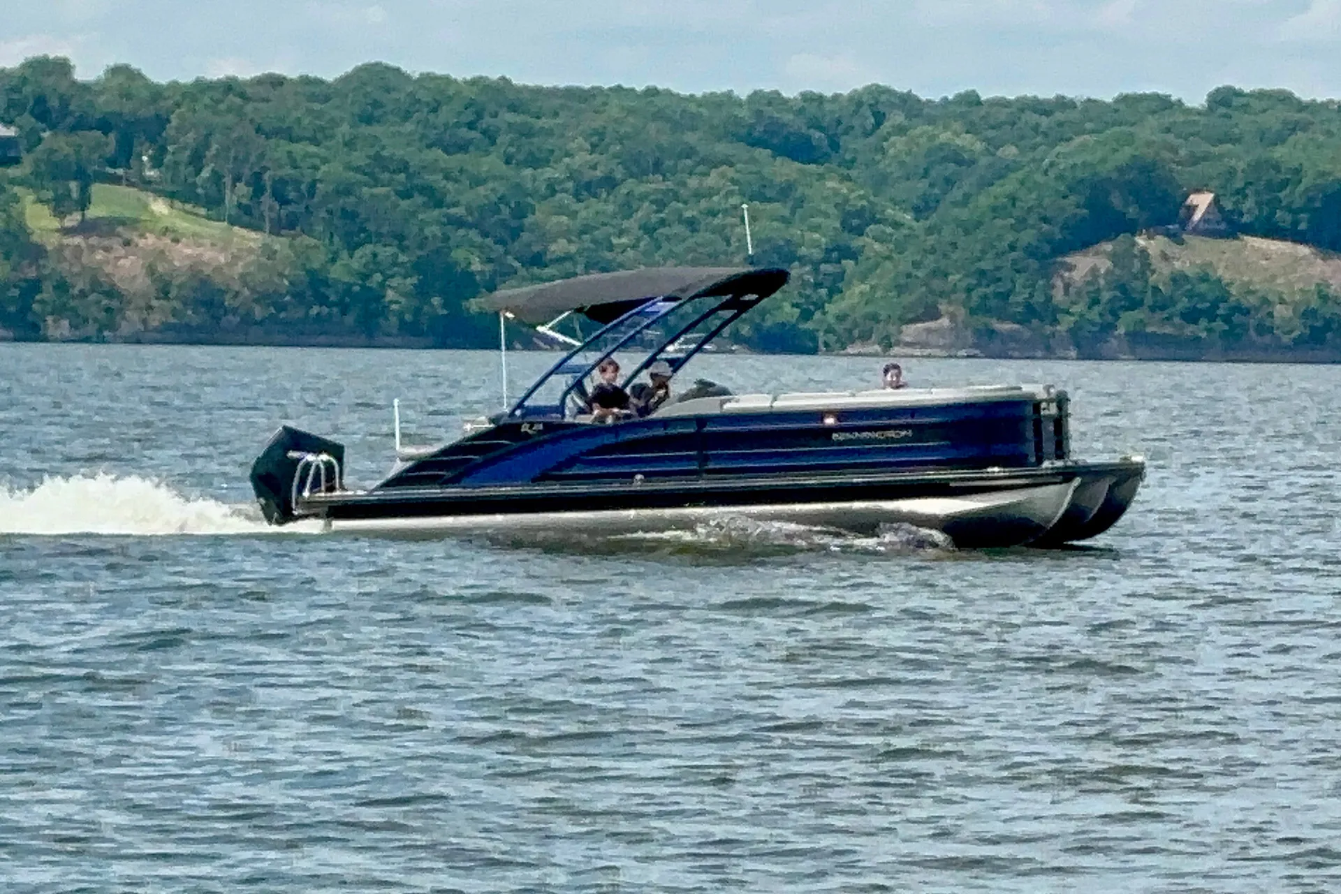 Blue Bennington pontoon boat with black canopy carrying three people on a lake near a forested shore.