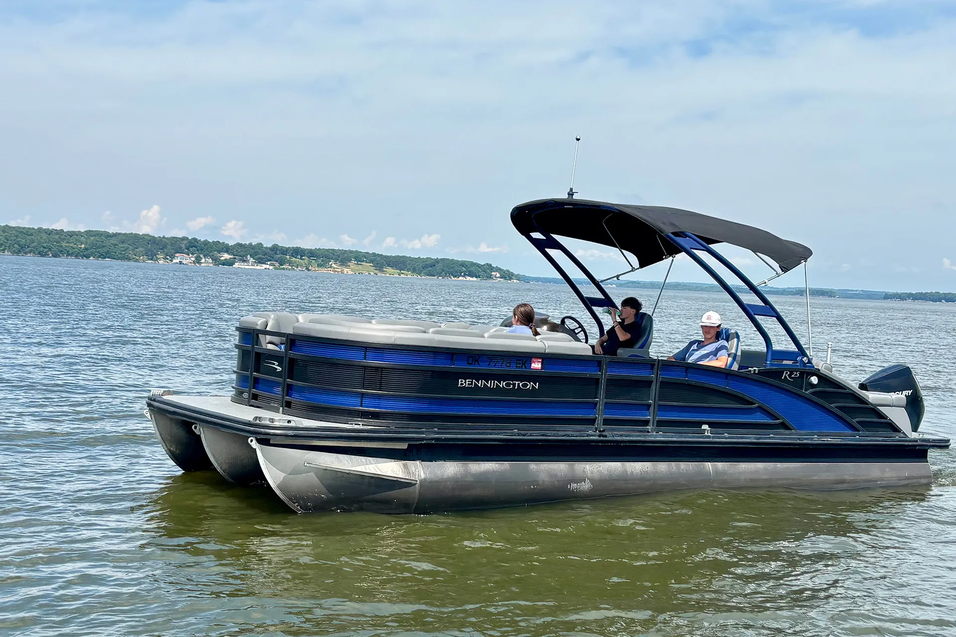 Blue and black Bennington pontoon boat with three people on calm lake water under a partly cloudy sky.