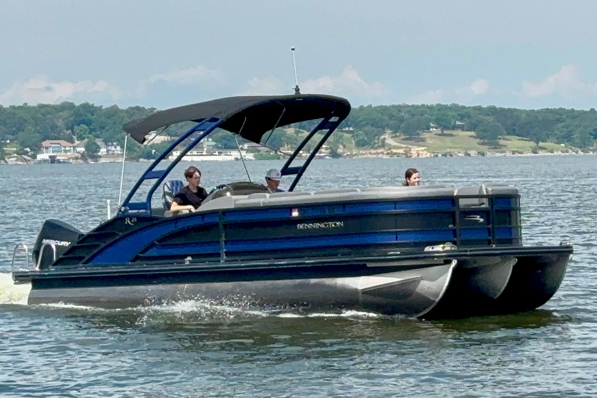 Blue Kennington pontoon boat with black canopy on water, carrying three people near shore.