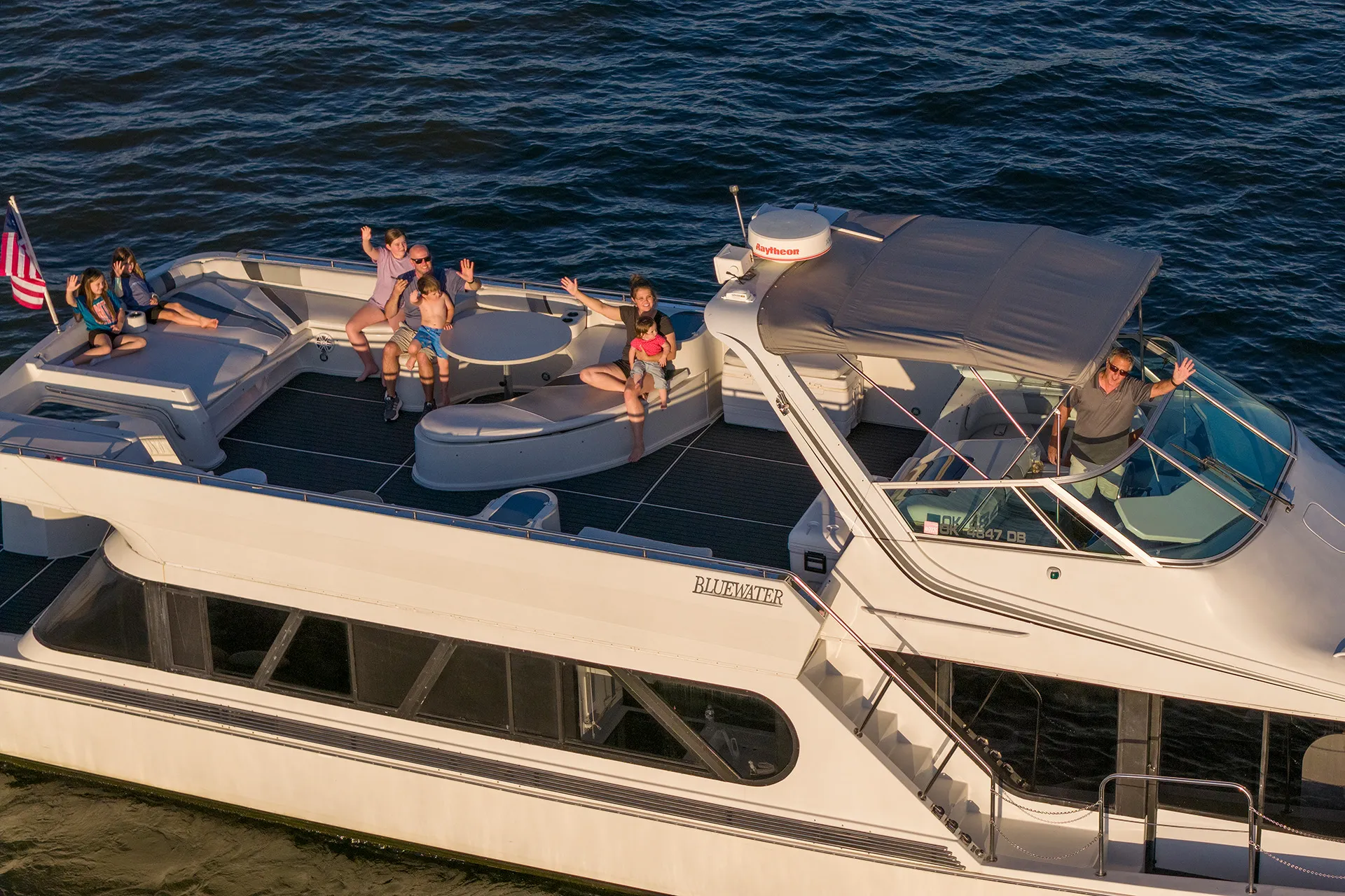 Family on a Bluewater boat waving, sitting on deck with water in the background.