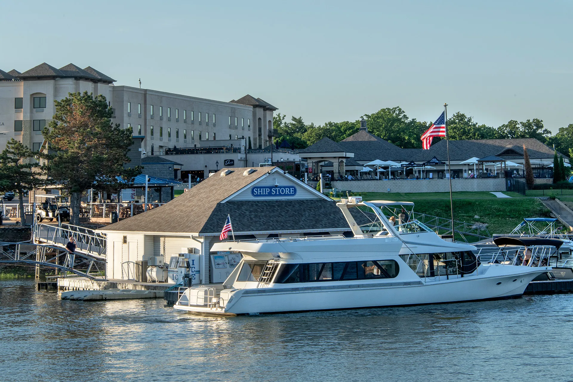 White boat docked next to a ship store by a waterfront with buildings and American flags nearby.