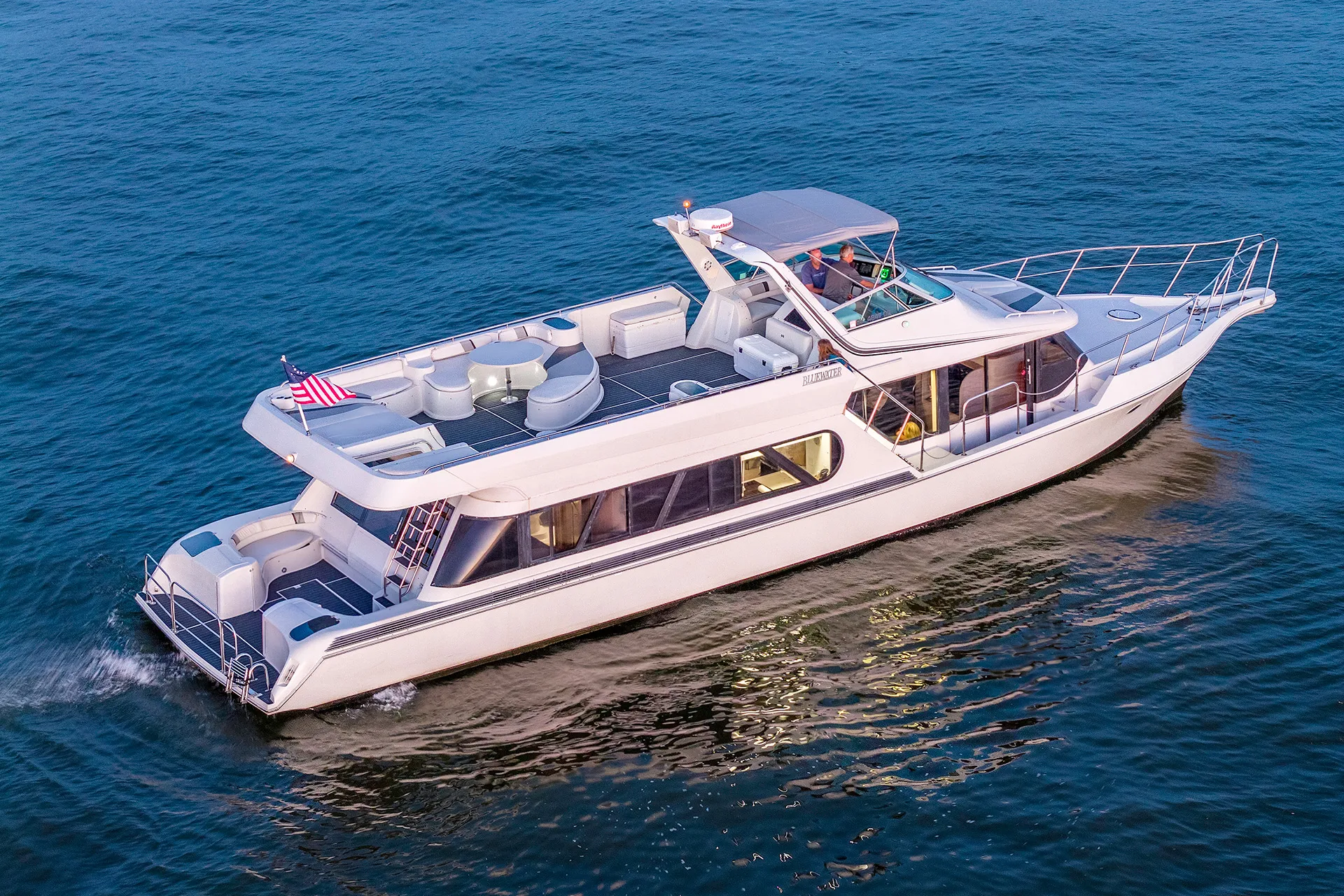 White luxury yacht cruising on calm blue water with people visible on deck and American flag at stern.