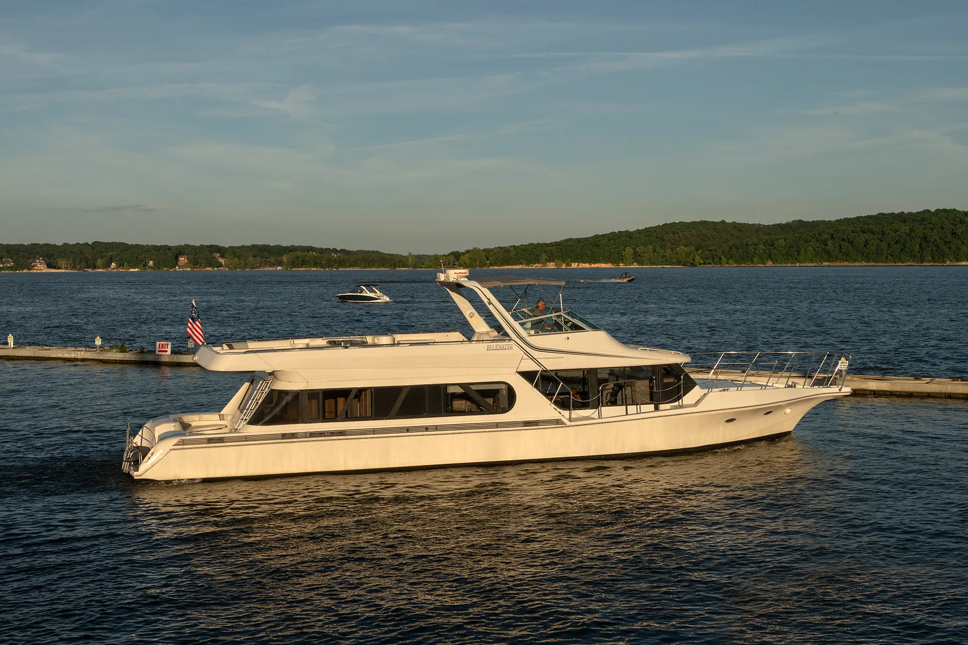 White motor yacht named Bluewater cruising on calm lake water with green shoreline in the background.