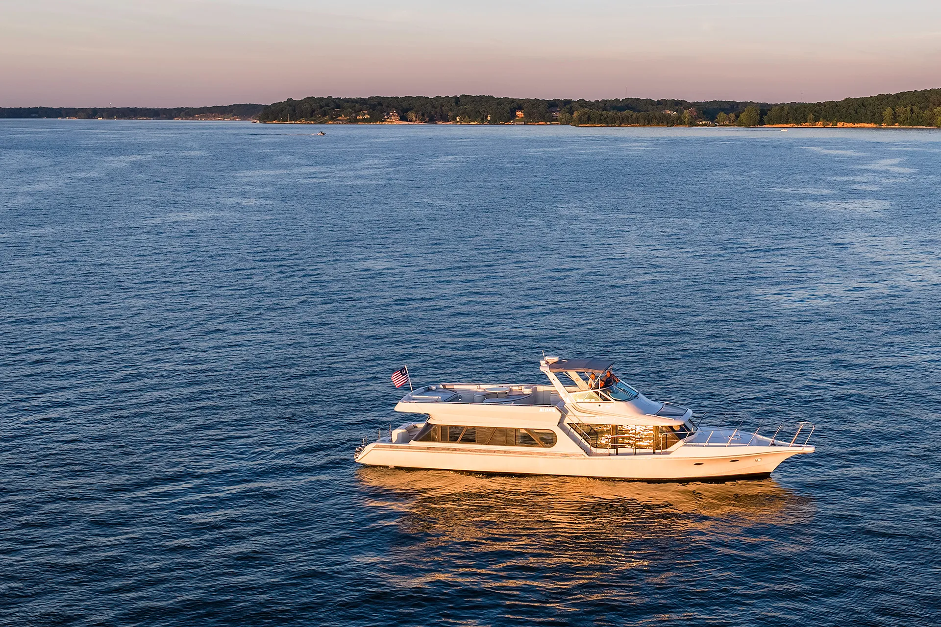 White luxury yacht cruising on calm blue water near a wooded shoreline at sunset.