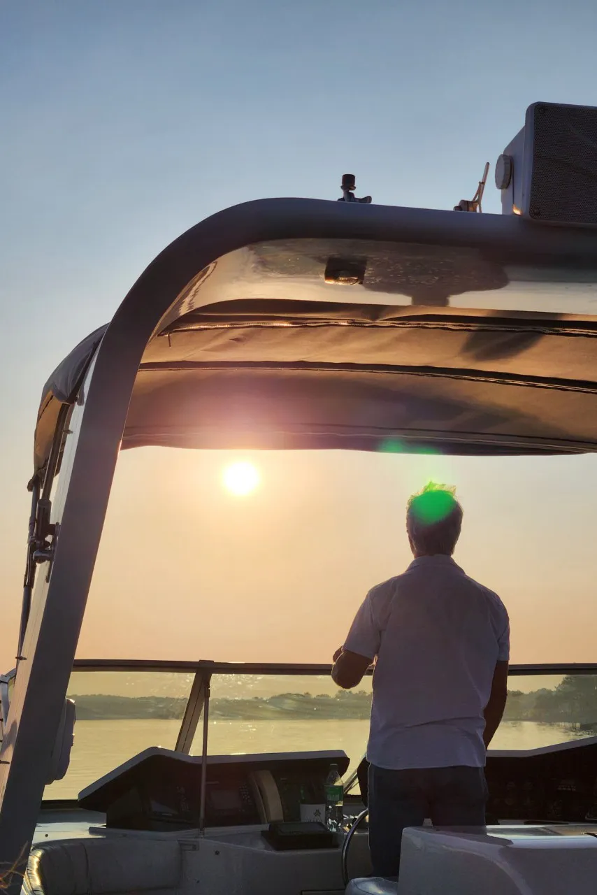 Person steering a boat at sunset with calm water and distant shoreline in the background.