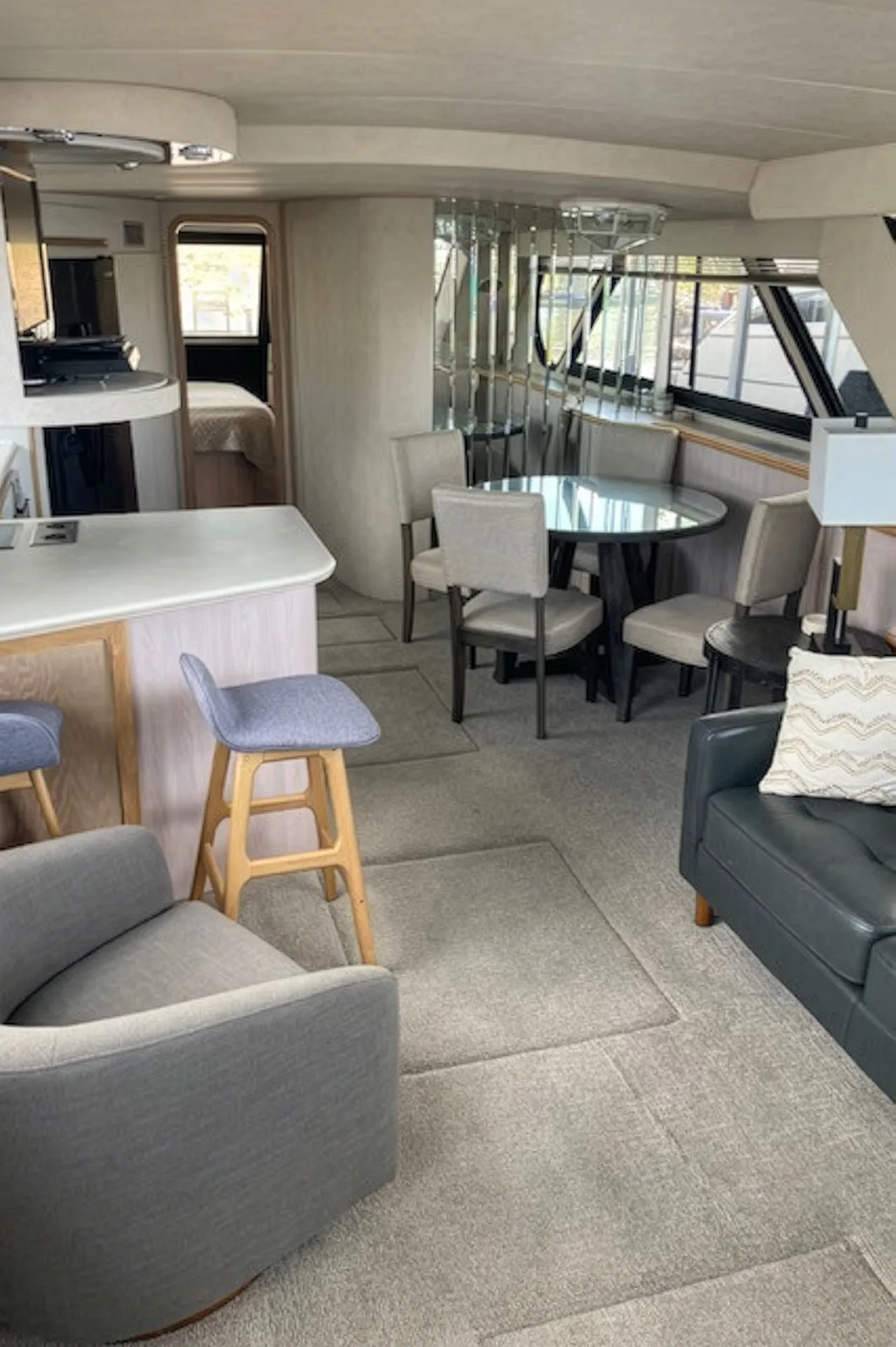 Bright yacht interior with dining table, gray chairs, bar stools, sofa, and a bed in the background.