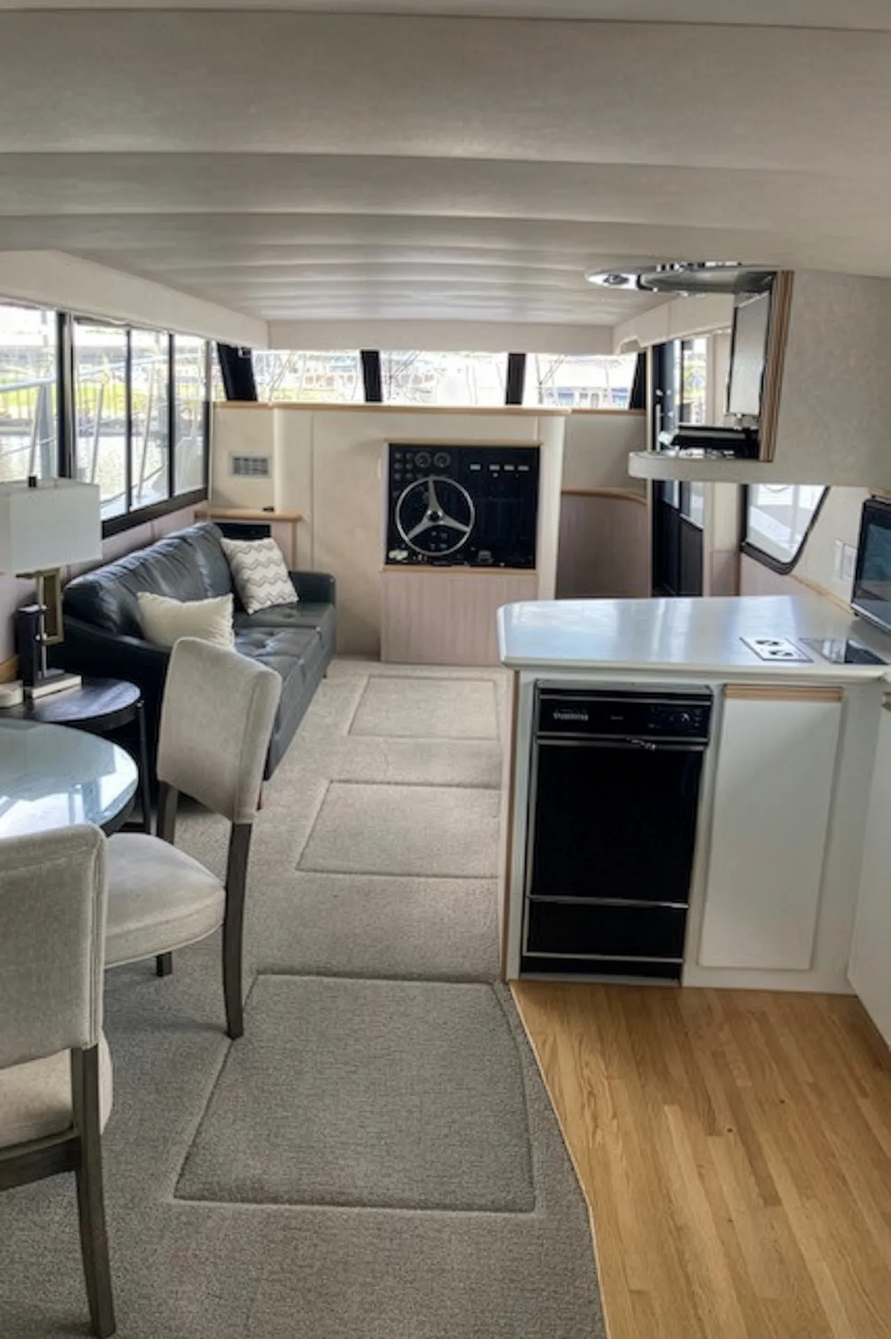 Interior of a boat with a black sofa, dining table, steering wheel, kitchen counter, and windows.