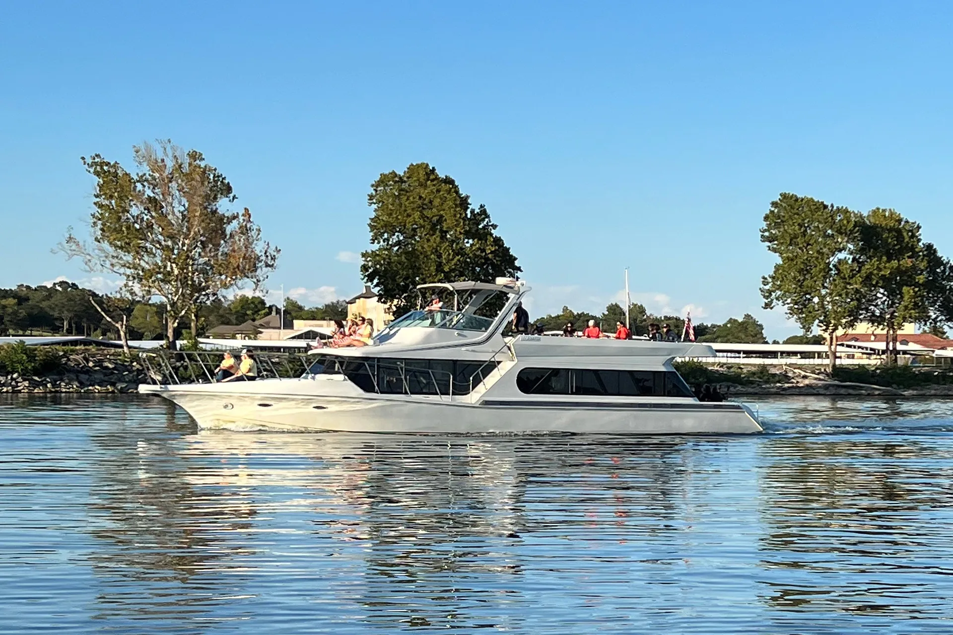 White yacht on calm water with people on deck and trees and buildings in the background.