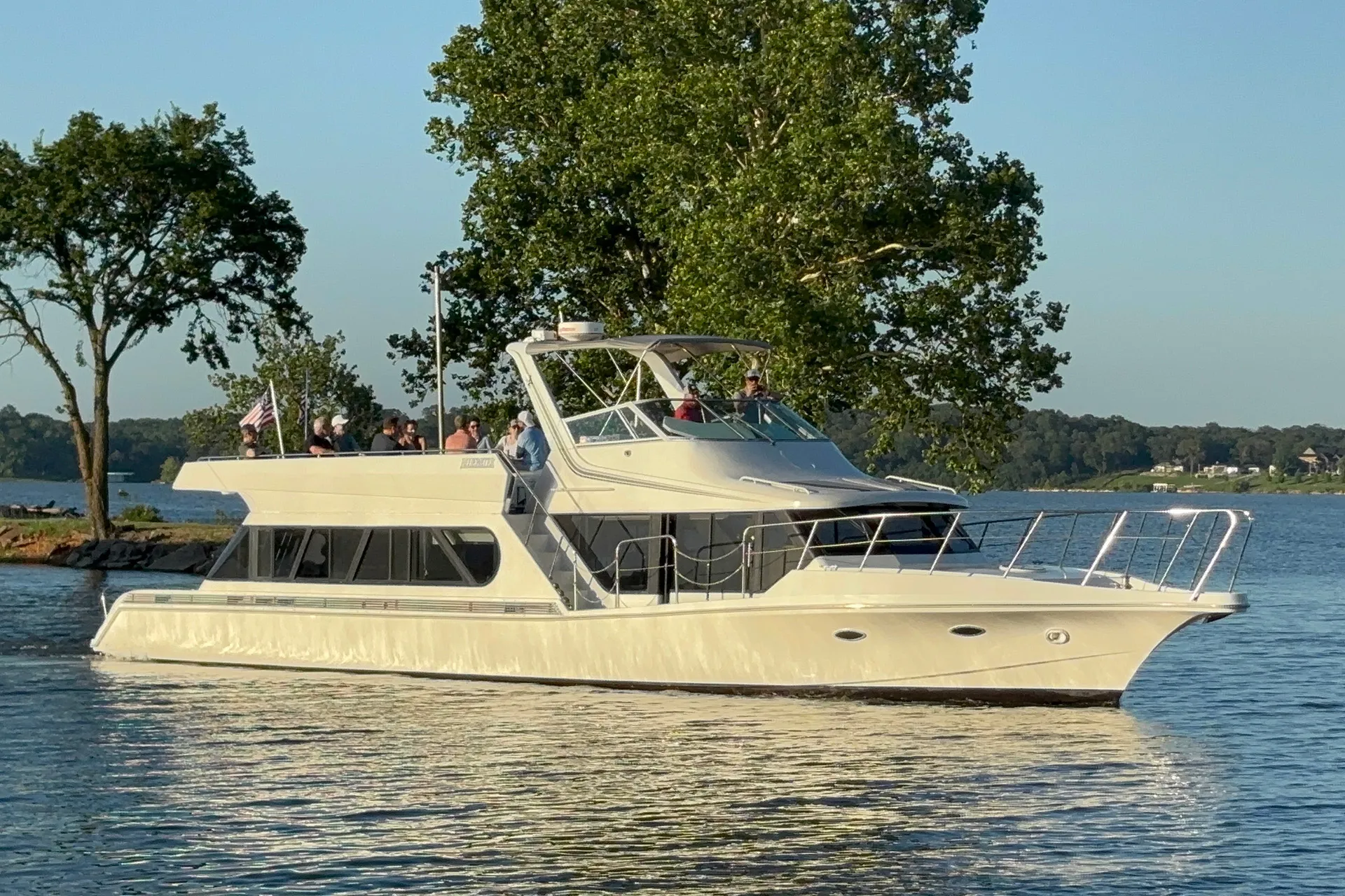 White yacht with people on deck cruising on calm water near green trees under clear sky.