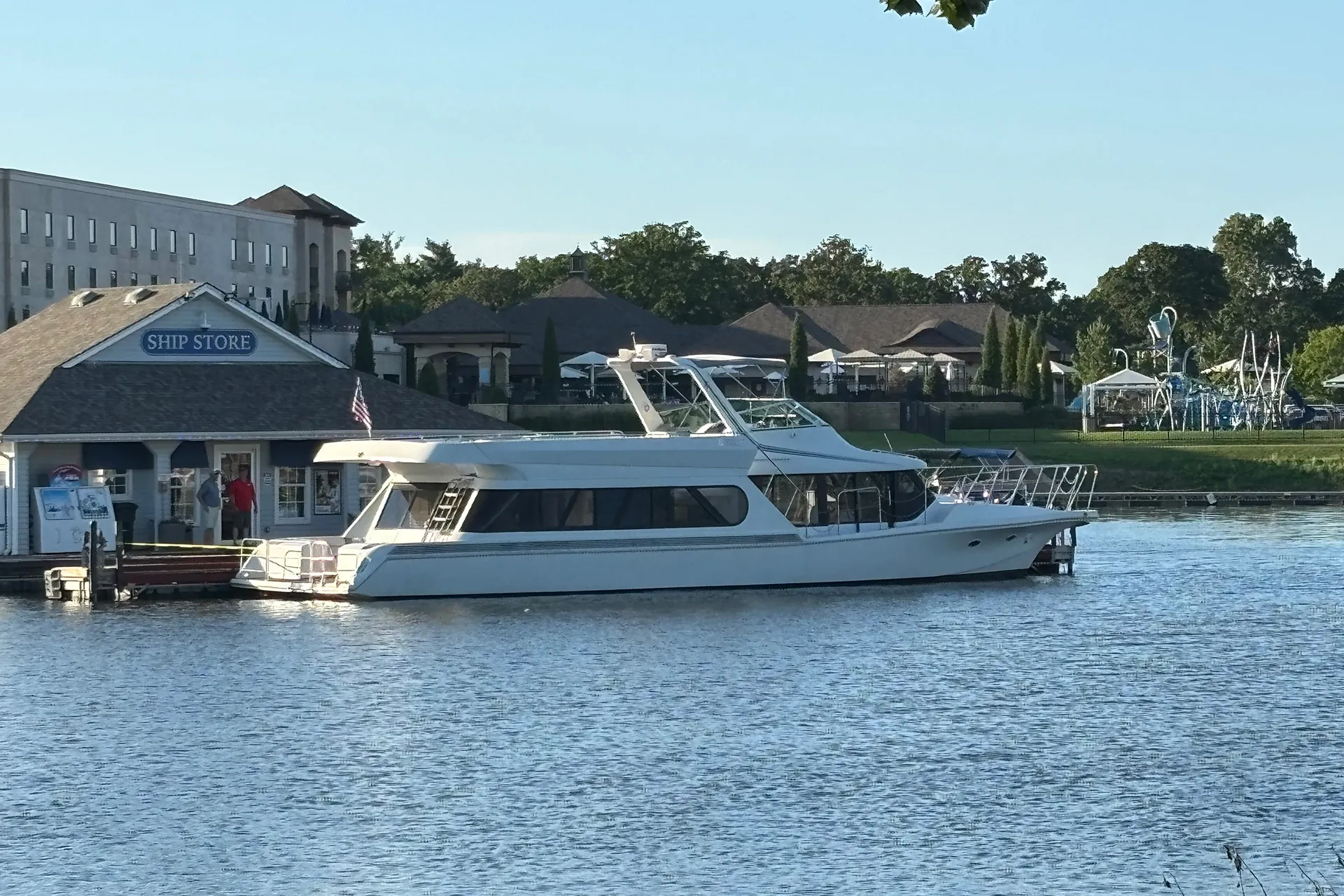 White yacht docked beside a ship store building on a calm river with greenery in the background.