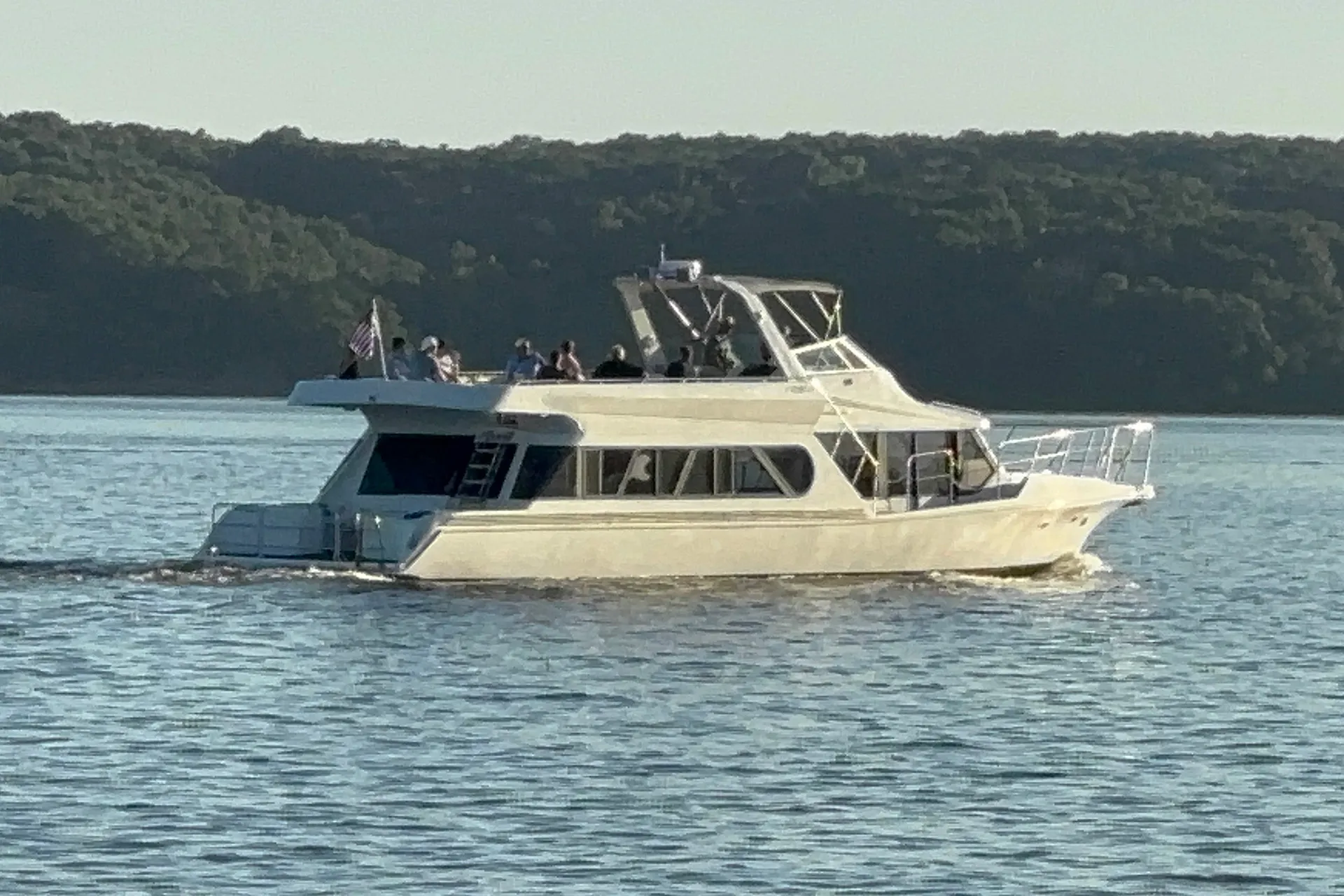 White yacht with people on upper deck cruising on calm water near forested shoreline.