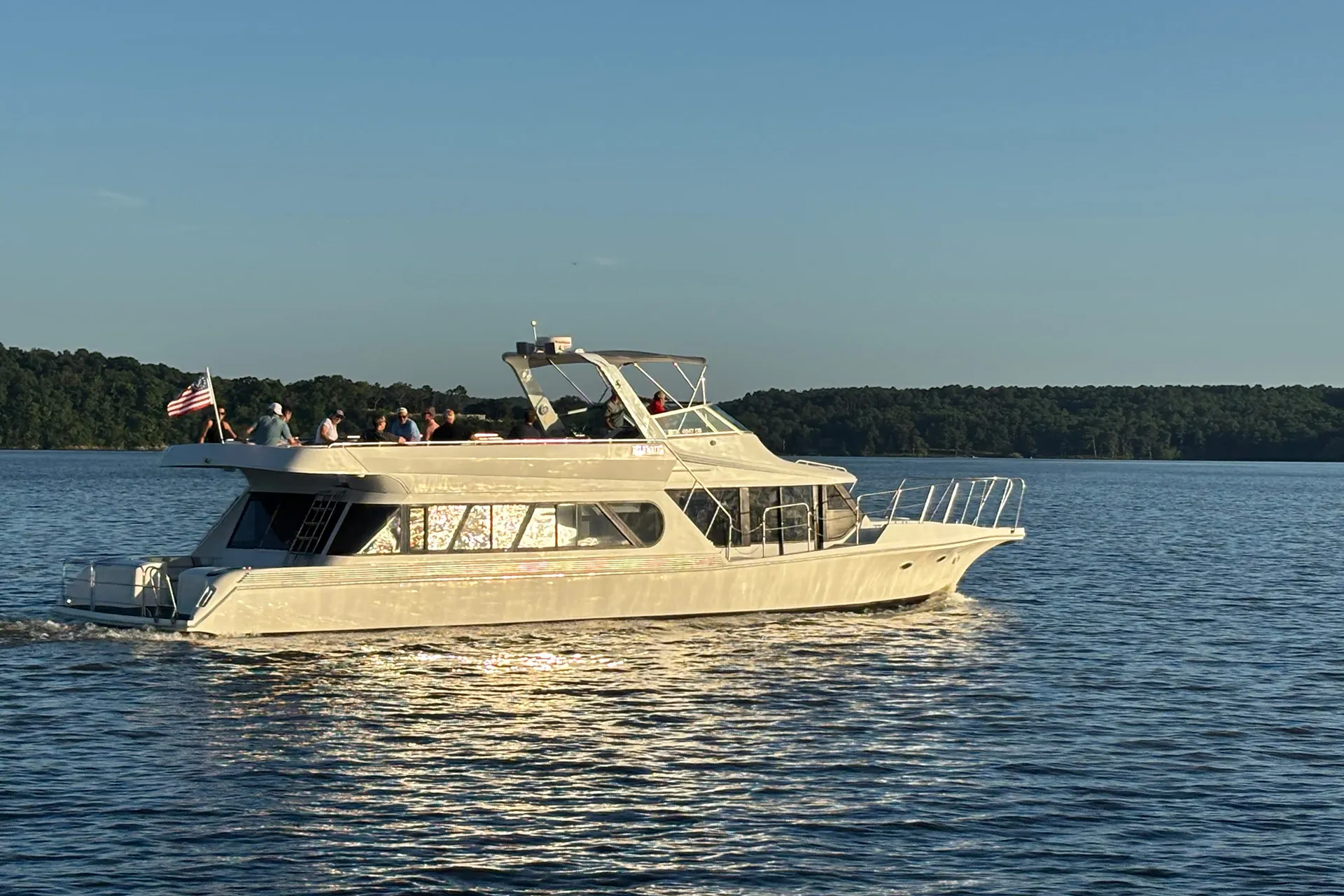 White yacht with people on deck cruising on calm water near forested shoreline under clear sky.