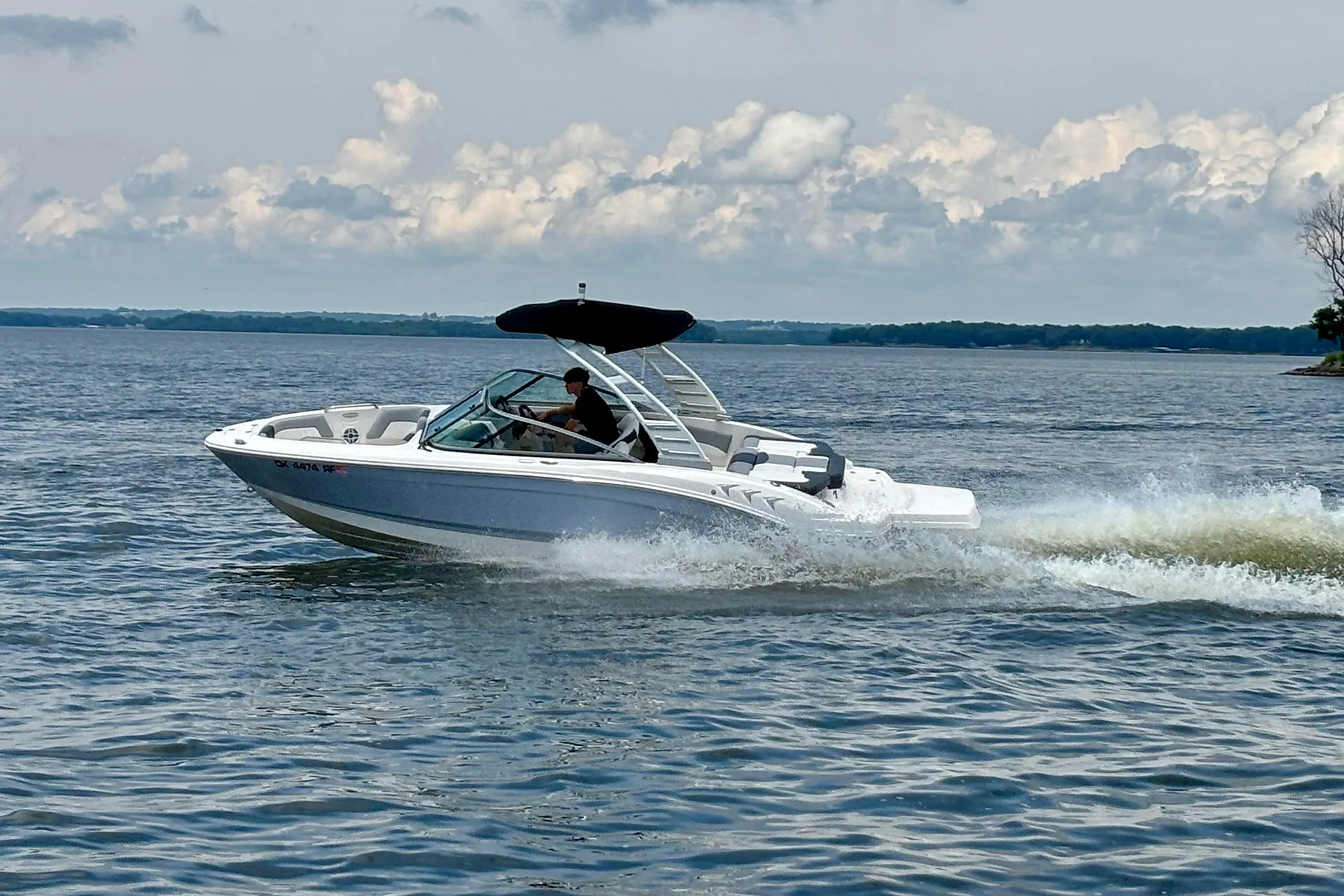 Person driving a white speedboat with black canopy on a lake under a cloudy sky.