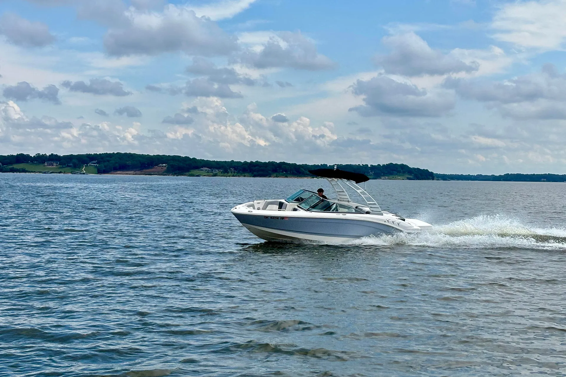 White motorboat speeding on calm lake water with a person steering under a partly cloudy sky.