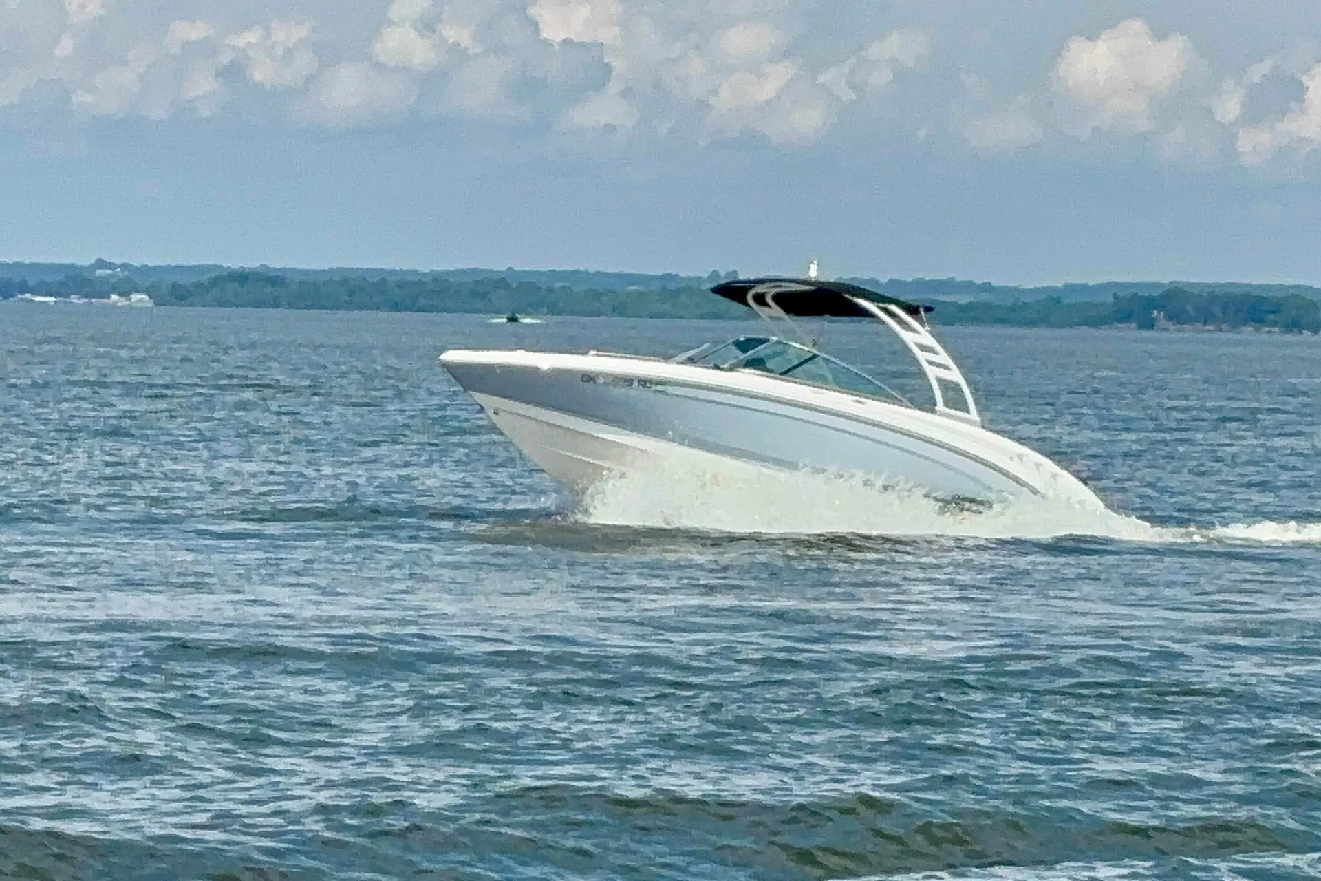 White motorboat speeding on water with waves and distant tree-lined shore under cloudy sky.
