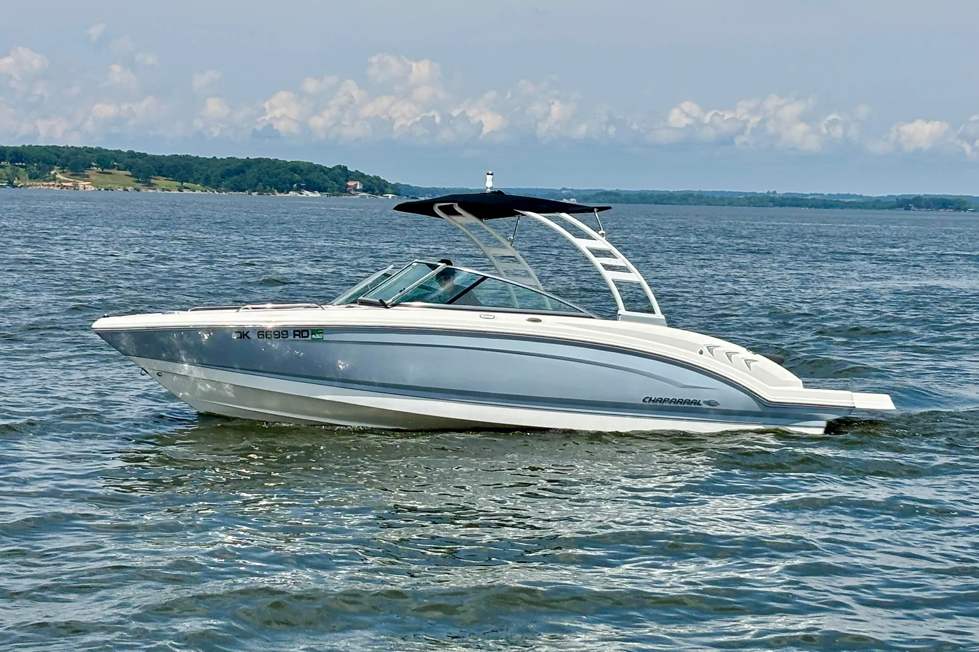 White Chaparral motorboat with black canopy cruising on calm water near a tree-lined shore.