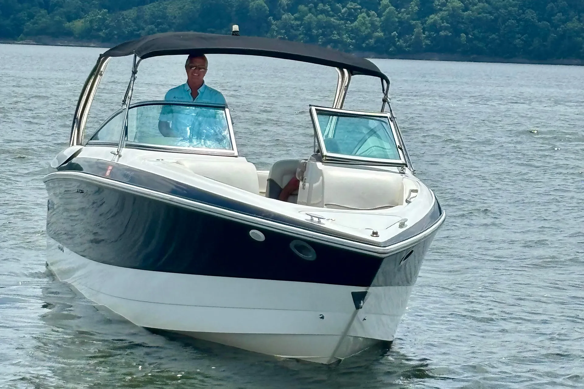Man steering a white and blue motorboat with black canopy on calm water near tree-lined shore.