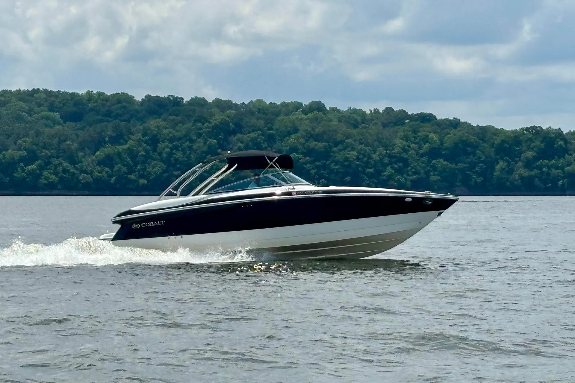 Black and white Cobalt motorboat speeding on lake water with forest and cloudy sky in background.