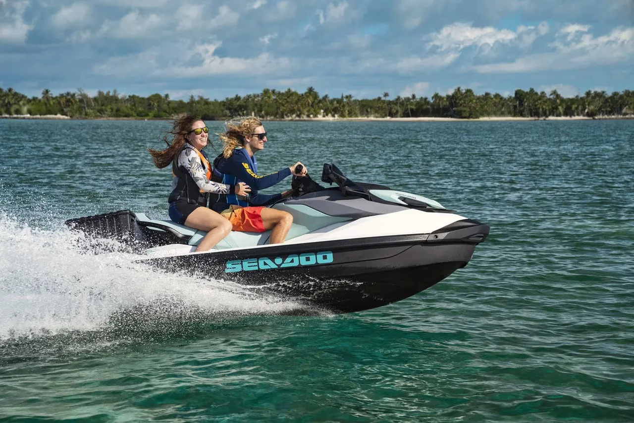 Two people riding a Sea-Doo jet ski on clear water near a tree-lined shore with cloudy sky.