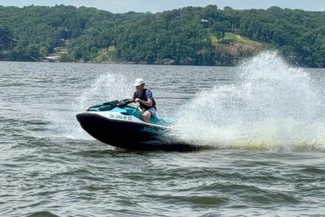 Person wearing helmet riding a Sea-Doo jet ski on a lake with trees in the background.
