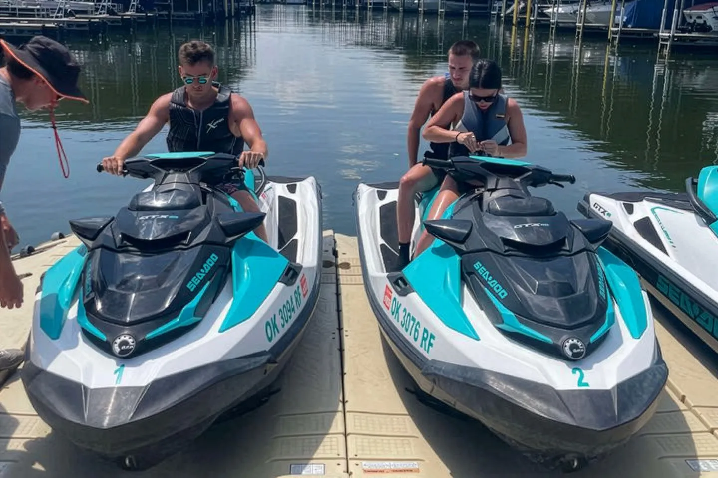 Two people on each of two parked Sea-Doo jet skis at a dock by the water.